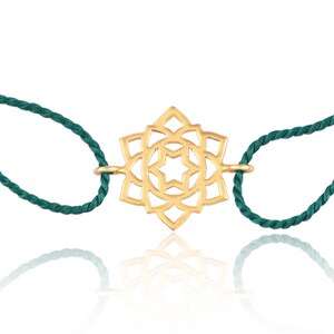 Puede incluir: Un brazalete de color dorado con un dije de mandala detallado. El brazalete está hecho de un cordón verde oscuro.