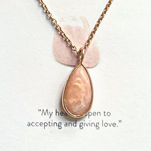 May include: A delicate gold chain necklace with a pink teardrop-shaped pendant. The pendant has a white marbled pattern and is set in a gold frame. The necklace is accompanied by a card with the text "My heart is open to accepting and giving love."