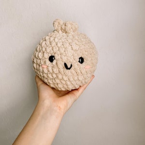 May include: A beige crocheted plush toy shaped like a dumpling with a smiling face. The toy has black eyes and a black mouth.