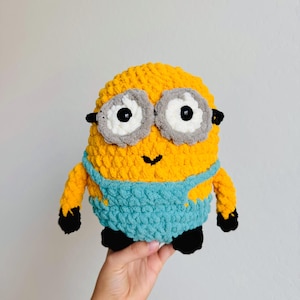 May include: A handmade crochet plush toy shaped like a yellow character with large white and grey goggles, black pupils, and a small black stitched smile. The figure wears blue overalls and has black hands and feet, crafted from soft, textured yarn.