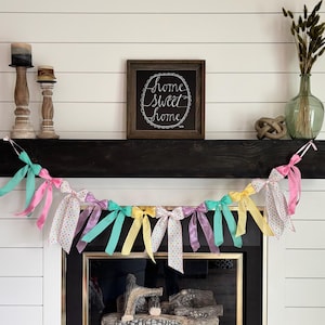 May include: A decorative garland with pastel ribbons in pink, teal, yellow, and lavender, hangs below a dark wooden mantel. The mantel also holds a framed sign that reads "Home Sweet Home."