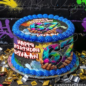 May include: A birthday cake with blue frosting and a colorful graphic design featuring the number 67 and the words "Happy Birthday RIVAAN". The cake is decorated with blue frosting and sits on a dark surface with confetti.