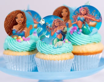 May include: Four cupcakes decorated with blue frosting and mermaid themed edible cupcake toppers. The toppers feature illustrations of mermaids with long flowing hair and tails.