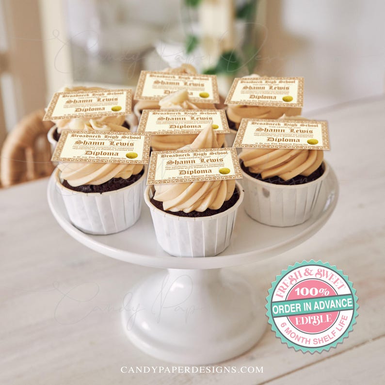 May include: Six chocolate cupcakes with vanilla frosting topped with edible images of graduation diplomas. The diplomas are printed with the text "Broadneck High School" and "Shann Lewis" with the word "Diploma" underneath. The cupcakes are displayed on a white cake stand.