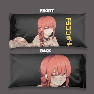 Puede incluir: Una funda de almohada rectangular negra con un personaje de estilo anime de pelo largo rosa y ojos naranjas. El frente muestra al personaje tumbado con texto japonés amarillo, mientras que el reverso muestra un primer plano del rostro.