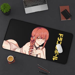 May include: Black rectangular mousepad featuring an anime-style illustration of a woman with red hair and yellow text in Japanese. A white computer mouse is on the right side of the mousepad. The mousepad is on a black desk.