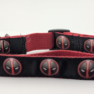 May include: Black and red dog collar featuring a repeating graphic of a character's mask. The collar has a black plastic buckle and a red fabric backing. The collar is approximately 1 inch wide.