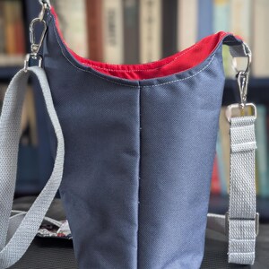 May include: A slate blue crossbody bag with a vibrant red interior lining. The bag features a textured exterior fabric, a vertical center seam, and a light gray adjustable woven strap attached with silver-toned metal swivel clasps and hardware.