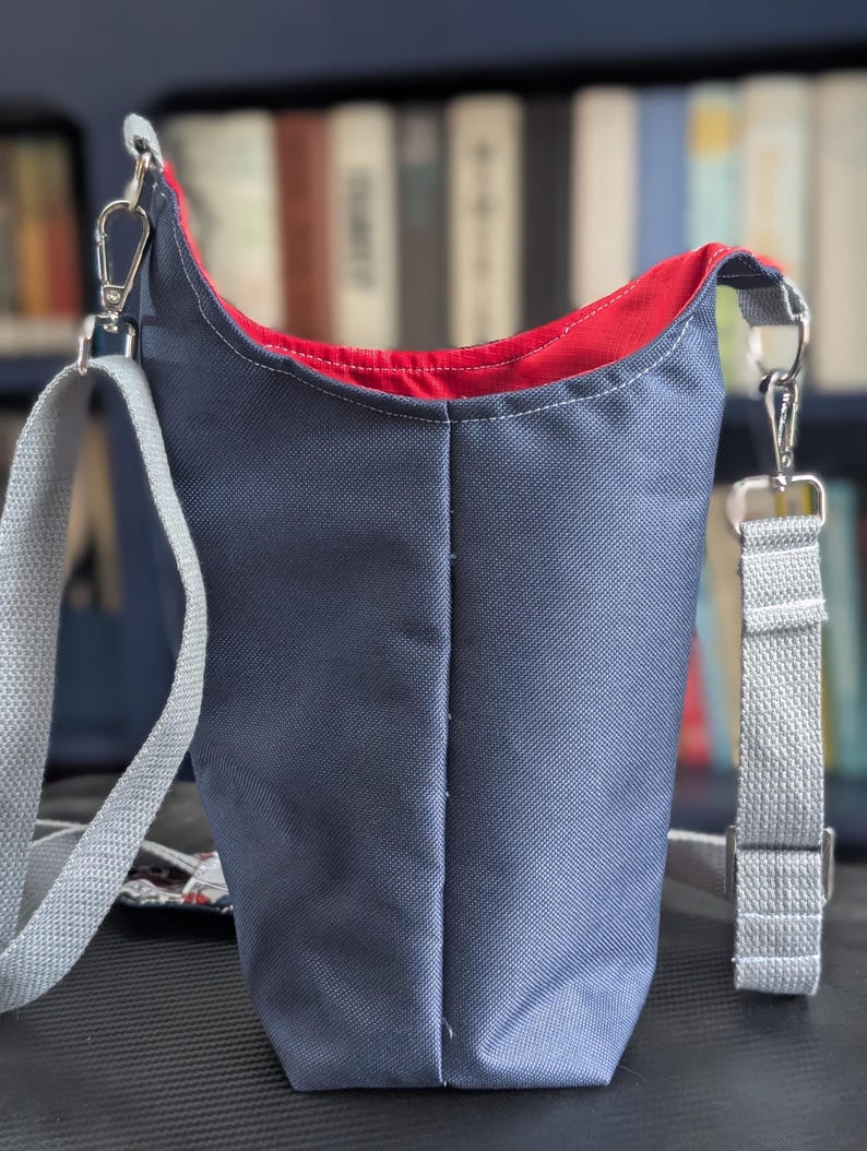 May include: A slate blue crossbody bag with a vibrant red interior lining. The bag features a textured exterior fabric, a vertical center seam, and a light gray adjustable woven strap attached with silver-toned metal swivel clasps and hardware.