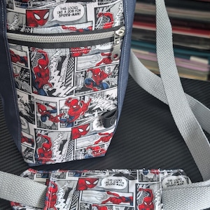 May include: A navy blue crossbody bag featuring a comic book print of Spider-Man with the text "THIS LOOKS LIKE A JOB FOR SPIDER-MAN". The bag includes a front zippered pocket, a red interior lining, and a matching padded shoulder strap.