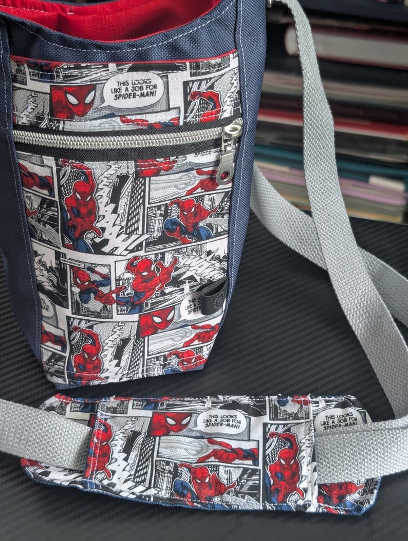 May include: A navy blue crossbody bag featuring a comic book print of Spider-Man with the text "THIS LOOKS LIKE A JOB FOR SPIDER-MAN". The bag includes a front zippered pocket, a red interior lining, and a matching padded shoulder strap.