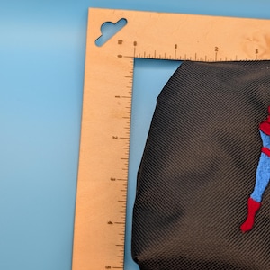 Puede incluir: Un estuche de tela negra con una figura bordada en un traje azul y rojo, colocado junto a una regla de madera en forma de L que muestra un ancho de aproximadamente 9 cm y una altura de aproximadamente 14 cm.