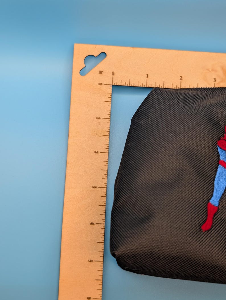 Puede incluir: Un estuche de tela negra con una figura bordada en un traje azul y rojo, colocado junto a una regla de madera en forma de L que muestra un ancho de aproximadamente 9 cm y una altura de aproximadamente 14 cm.