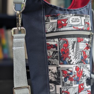 May include: A navy blue crossbody bag featuring a front pocket with a black and white comic book print of Spider-Man in red and blue. The bag has a red interior lining, a grey adjustable strap with a silver metal clasp, and a silver zipper closure.