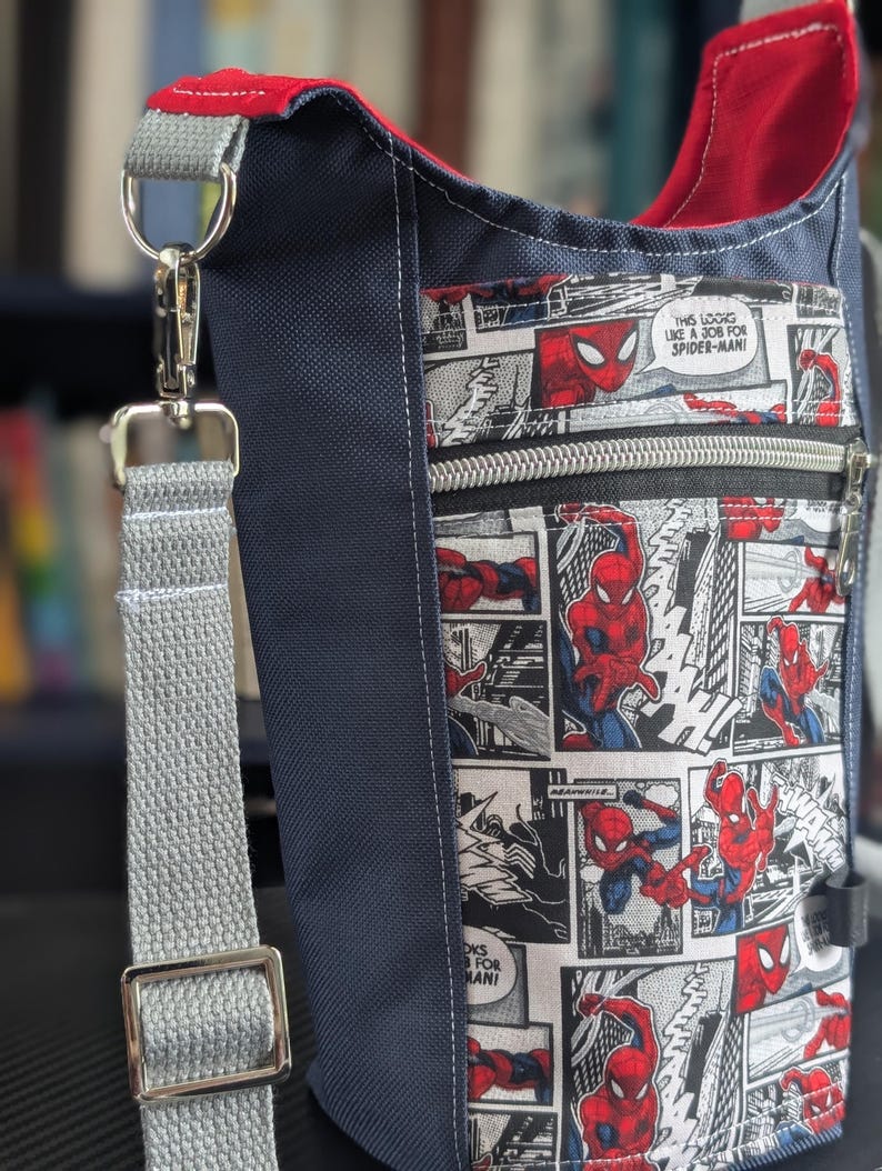 May include: A navy blue crossbody bag featuring a front pocket with a black and white comic book print of Spider-Man in red and blue. The bag has a red interior lining, a grey adjustable strap with a silver metal clasp, and a silver zipper closure.