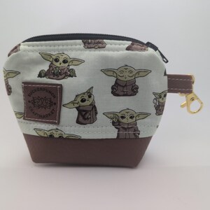 May include: A small, square coin purse with a zipper closure. The top portion is light blue with a repeating pattern of a cartoon character. The bottom is brown. A brown leather strap and gold-colored clasp are attached to the side. A brown patch with text is on the front.