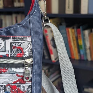 May include: A crossbody bag featuring a comic book print of Spider-Man in red and blue on a white background. The bag has a dark blue textured fabric side panel, a red interior lining, a silver metal zipper, and a light gray adjustable webbing strap.