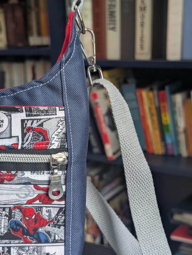 May include: A crossbody bag featuring a comic book print of Spider-Man in red and blue on a white background. The bag has a dark blue textured fabric side panel, a red interior lining, a silver metal zipper, and a light gray adjustable webbing strap.