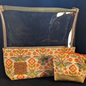 Pode incluir: Uma bolsa grande de vinil transparente com zíper verde e uma bolsa menor correspondente, ambas com base de tecido creme com um padrão repetitivo de flores laranja e besouros verdes. Um emblema quadrado de couro marrom com o texto Suckerpunch.