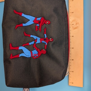 Puede incluir: Un estuche negro con cremallera que presenta un bordado de tres figuras con trajes rojos y azules se&ntilde;al&aacute;ndose entre s&iacute;. Una regla de madera es visible en los bordes superior y derecho, mostrando una anchura de unos 20 cm.