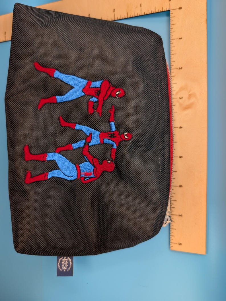 Puede incluir: Un estuche negro con cremallera que presenta un bordado de tres figuras con trajes rojos y azules se&ntilde;al&aacute;ndose entre s&iacute;. Una regla de madera es visible en los bordes superior y derecho, mostrando una anchura de unos 20 cm.
