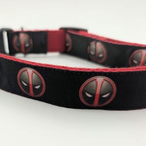 May include: Black dog collar with a red trim and repeating Deadpool mask design. The collar features a black buckle and a red fabric section. The Deadpool mask is a stylized oval with white eyes and a red vertical line.
