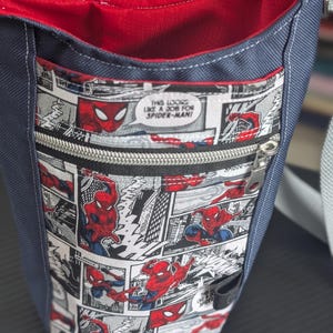 May include: A navy blue crossbody bag with a red interior lining and a front zippered pocket featuring a black and white comic book print of Spider-Man. The comic panels include the text, "THIS LOOKS LIKE A JOB FOR SPIDER-MAN!" and a silver zipper.