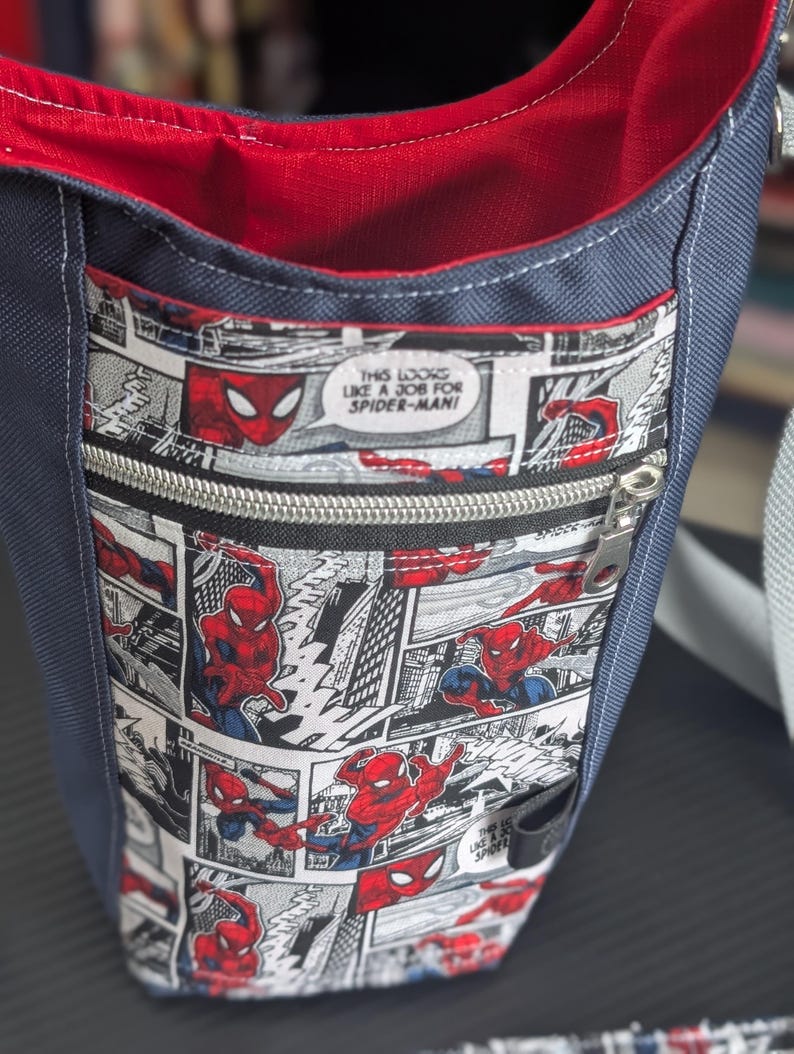 May include: A navy blue crossbody bag with a red interior lining and a front zippered pocket featuring a black and white comic book print of Spider-Man. The comic panels include the text, "THIS LOOKS LIKE A JOB FOR SPIDER-MAN!" and a silver zipper.