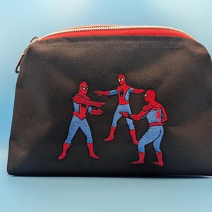 Puede incluir: Un estuche negro con cremallera que presenta un bordado de tres figuras de Spider-Man en la ic&oacute;nica pose del meme se&ntilde;al&aacute;ndose. El estuche tiene un cierre de cremallera rojo en el borde superior y una peque&ntilde;a etiqueta de tela azul oscuro.