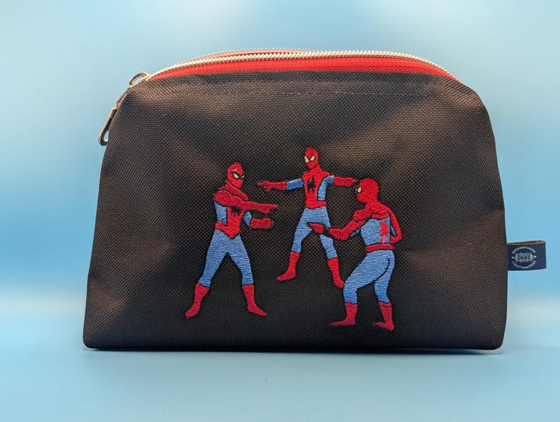 Puede incluir: Un estuche negro con cremallera que presenta un bordado de tres figuras de Spider-Man en la ic&oacute;nica pose del meme se&ntilde;al&aacute;ndose. El estuche tiene un cierre de cremallera rojo en el borde superior y una peque&ntilde;a etiqueta de tela azul oscuro.