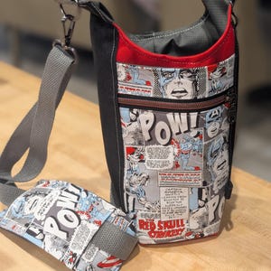 May include: A black and red water bottle holder and matching phone holder featuring a comic book design with Captain America graphics and the text "POW!" and "Red Skull Strikes!". The bag has a gray strap and a zippered pocket.