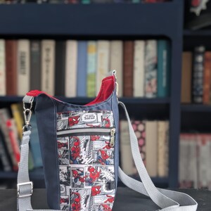 May include: A crossbody bag featuring a comic book print of Spider-Man in red and blue on a white background. The bag has a solid navy blue exterior, a red interior lining, a front zippered pocket, and an adjustable light gray woven shoulder strap.