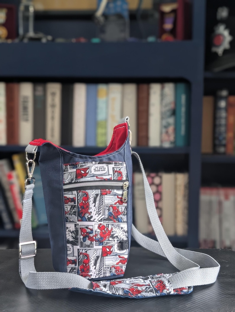 May include: A crossbody bag featuring a comic book print of Spider-Man in red and blue on a white background. The bag has a solid navy blue exterior, a red interior lining, a front zippered pocket, and an adjustable light gray woven shoulder strap.