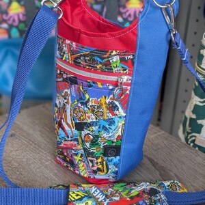 May include: A blue and red crossbody bag with a comic book print. The bag features a zippered pocket and a blue adjustable strap. The interior is red, and the exterior is a collage of comic book panels.
