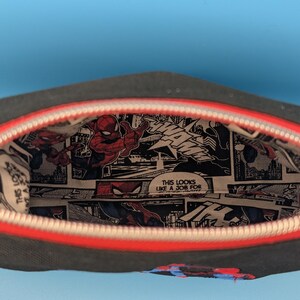 Puede incluir: Un estuche negro con cremallera y borde rojo, con un forro interior estampado con un patr&oacute;n de c&oacute;mic de Spider-Man, que incluye el texto "THIS LOOKS LIKE A JOB FOR" y "WHAM" en paneles en blanco y negro con detalles en rojo y azul.
