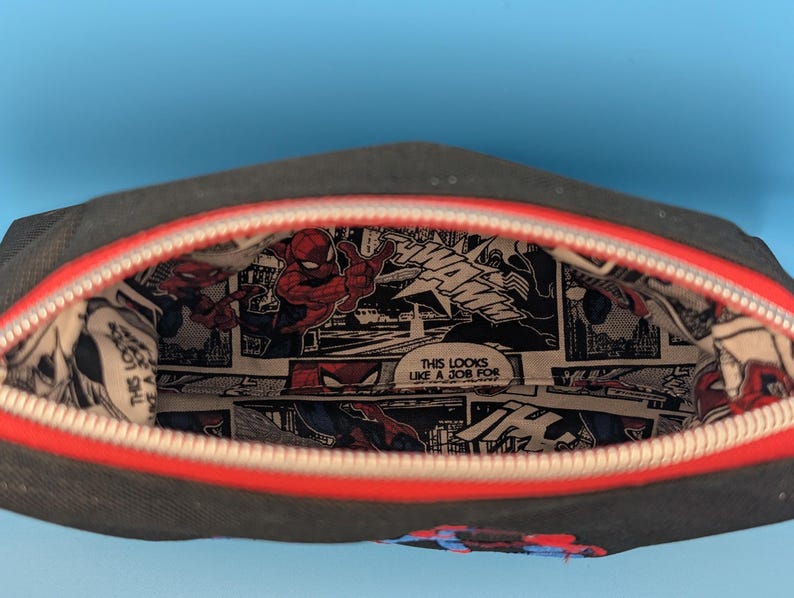 Puede incluir: Un estuche negro con cremallera y borde rojo, con un forro interior estampado con un patr&oacute;n de c&oacute;mic de Spider-Man, que incluye el texto "THIS LOOKS LIKE A JOB FOR" y "WHAM" en paneles en blanco y negro con detalles en rojo y azul.