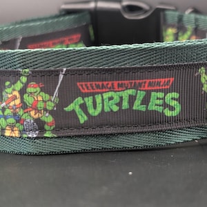 May include: Green dog collar featuring the Teenage Mutant Ninja Turtles logo and characters. The collar has a black band with the TMNT logo and images of the turtles. The collar is made of a durable material and has a plastic buckle.
