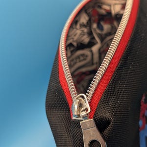 Puede incluir: Un estuche negro con cremallera que presenta un tirador de metal plateado y un borde de tela roja a lo largo de la abertura. El forro interior est&aacute; decorado con un estampado gr&aacute;fico en blanco y negro estilo c&oacute;mic con ilustraciones.