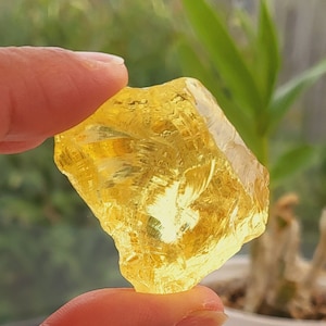 Puede incluir: Un gran cristal amarillo claro con una superficie rugosa y facetada. El cristal está siendo sostenido por una mano.