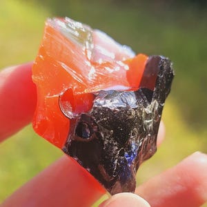 Puede incluir: Una mano sostiene un trozo de obsidiana en bruto pulida. La piedra es una mezcla de naranja y negro, con una superficie vítrea y reflectante. El fondo es verde borroso.