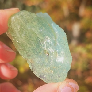 Puede incluir: Una gema translúcida de color verde claro sostenida en una mano. La piedra en bruto tiene una forma irregular y una textura natural. El fondo está borroso, con toques de follaje verde y marrón.