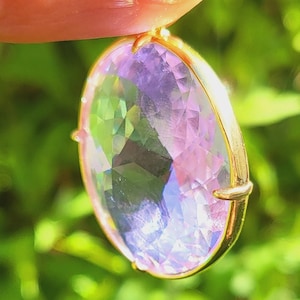 Puede incluir: Primer plano de un colgante de piedra preciosa púrpura con engaste de oro. El colgante está siendo sostenido por un dedo con esmalte de uñas rojo.