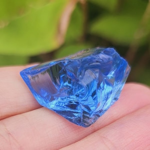 Könnte beinhalten: Ein facettiertes, unregelmäßig geformtes Stück blaues Glas. Das Glas hat ein kristallines Aussehen und wird in einer Hand gehalten. Der Hintergrund ist verschwommenes grünes Laub.