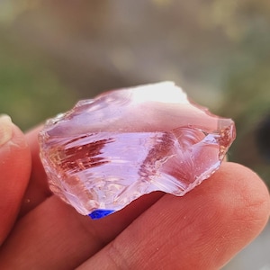 Puede incluir: Un trozo de vidrio translúcido rosa-púrpura con una forma rugosa e irregular. El vidrio tiene un ligero tinte azul en un borde. La imagen es un primer plano, que destaca la textura y el color del vidrio.