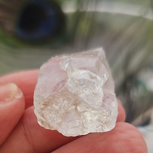 Puede incluir: Un cristal transparente y facetado con un ligero tono rosado. El cristal se sostiene en una mano.