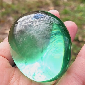 Puede incluir: Una esfera de vidrio lisa y verde con un patrón de remolino en su interior. La esfera está siendo sostenida en una mano.