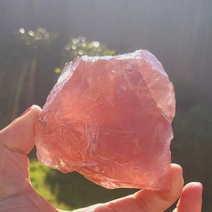May include: A large, pink, translucent crystal with a rough, natural texture. The crystal is being held in a hand against a blurred background of green grass and a blue sky.