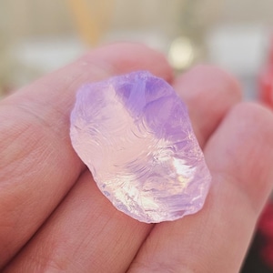May include: A translucent, light purple gemstone held in a hand. The stone has a smooth, polished appearance with subtle internal patterns. The color transitions from a pale pink to a deeper lavender hue.