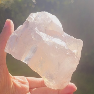 May include: A large, clear, translucent, pink quartz crystal with a rough, natural texture. The crystal is being held in a hand against a blurred background of green grass and trees.