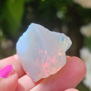 Puede incluir: Un cristal de ópalo blanco con colores arcoíris iridiscentes. El cristal es áspero y tiene un aspecto natural sin pulir.